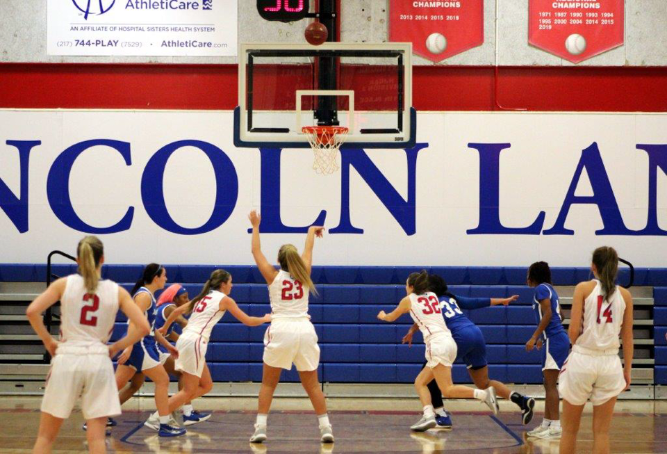 A group of women's basketball players gather under a hoop as a player shoots the ball. 