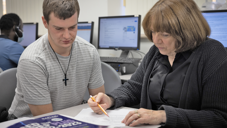 A student looks down at a piece of paper that a teacher is pointing to with a pencil.