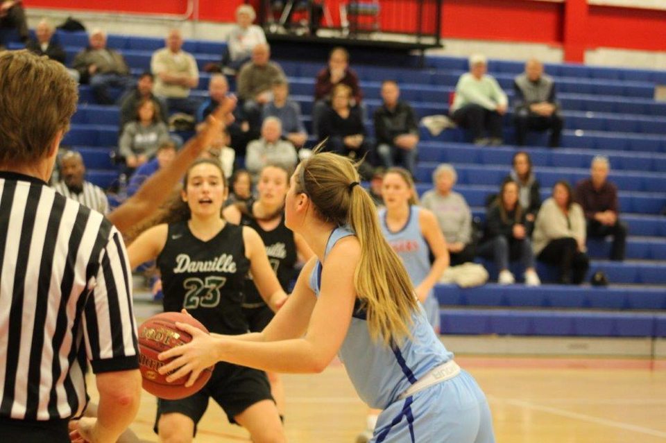 An LLCC women's basketball player in a blue uniform prepares to pass the ball while being guarded by a player from the opposing team. A crowd looks on from the bleachers in the background. 