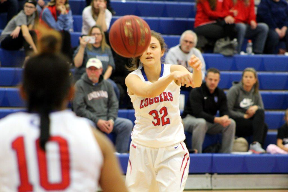 An LLCC women's basketball player in a white uniform throws a ball towards a fellow player while a crowd sits behind her in the bleachers. 