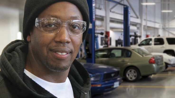 A student smiles; in the background are cars in an auto lab.