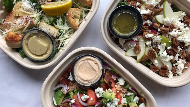 Three to-go containers with salads and individual cups of salad dressing.