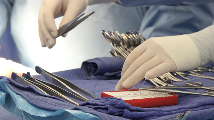 Two hands wearing white surgical gloves reach for surgical tools on a tray.