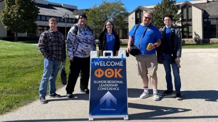 LLCC Phi Theta Kappa officers stand outside by a folding sign that says "welcome Illinois regional leadership conference."
