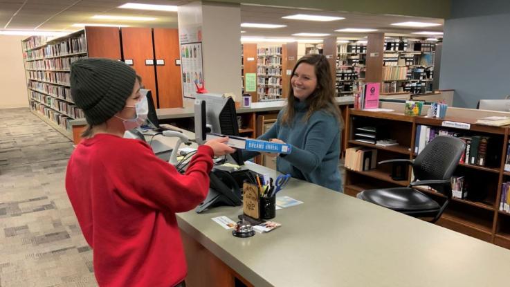 A student hands a book over the counter to a librarian to check out.