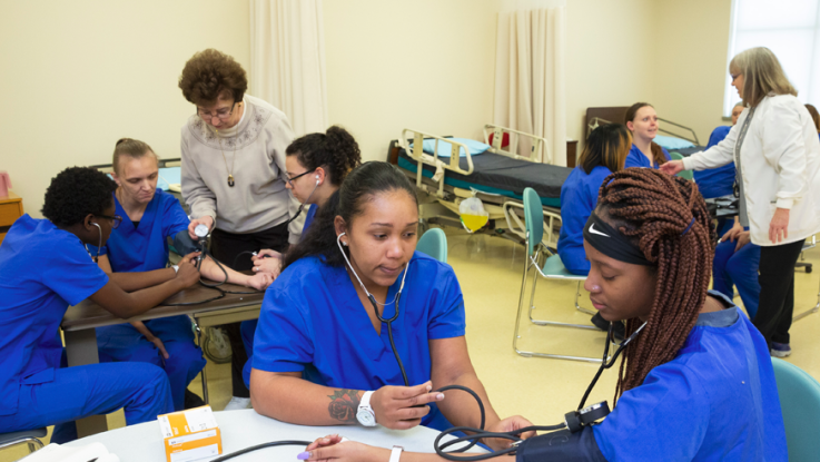 Nine students in scrubs learning from two instructors about how to take blood pressure readings