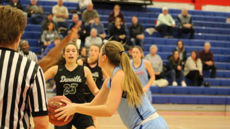 An LLCC women's basketball player in a blue uniform prepares to pass the ball while being guarded by a player from the opposing team. A crowd looks on from the bleachers in the background. 