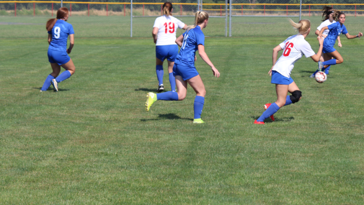 Women in soccer uniforms run across the field kicking the soccer ball. 