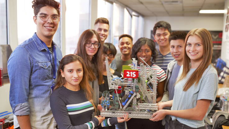 Young students stand in a circle and smile while holding a robotics project.