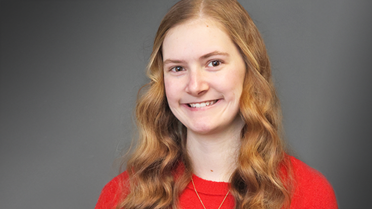 A female with red hair wears a red shirt and silver cross necklace and smiles for a headshot photo.