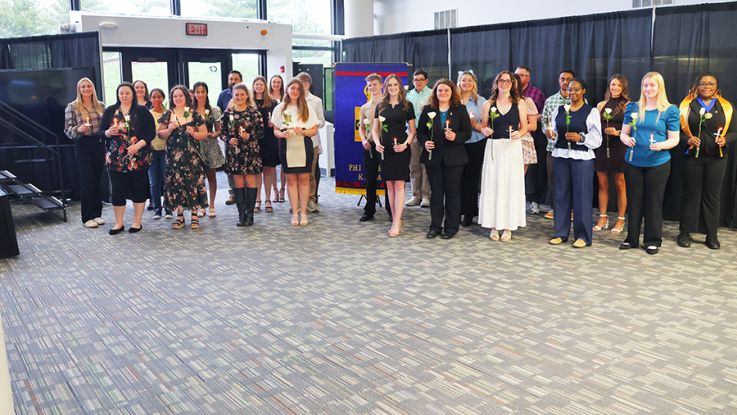 Large group of people stand in a lobby holding lit candles during an induction ceremony, with a banner and black curtain backdrop behind them.