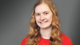A female with red hair wears a red shirt and silver cross necklace and smiles for a headshot photo.