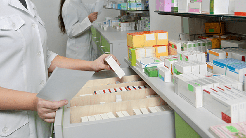 pharmacy technician holding medications in front of a shelf stocked with various medications