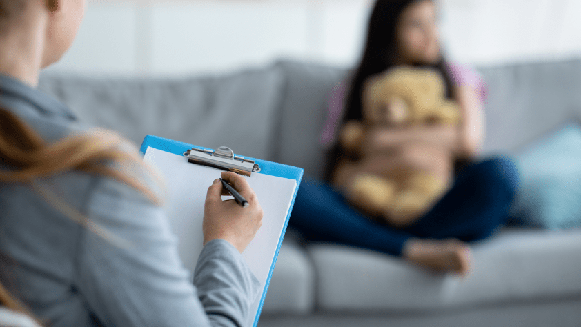 counselor holding clipboard and pen taking notes while in a session with a child seated on a couch hugging a stuffed animal