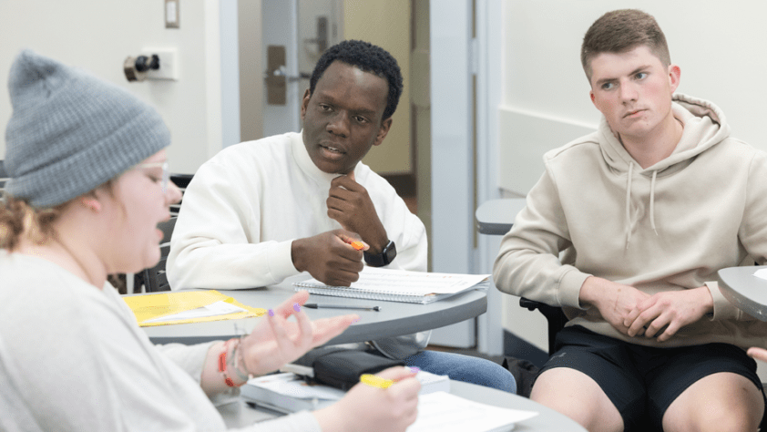 three students at their desks in a discussion