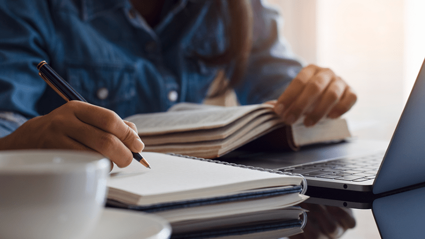 person with book and laptop open, starting to write in notebook