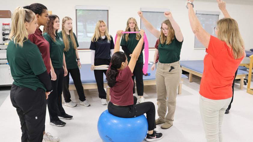 instructor and students in classroom assistant a student doing band shoulder stretches while sitting on big ball