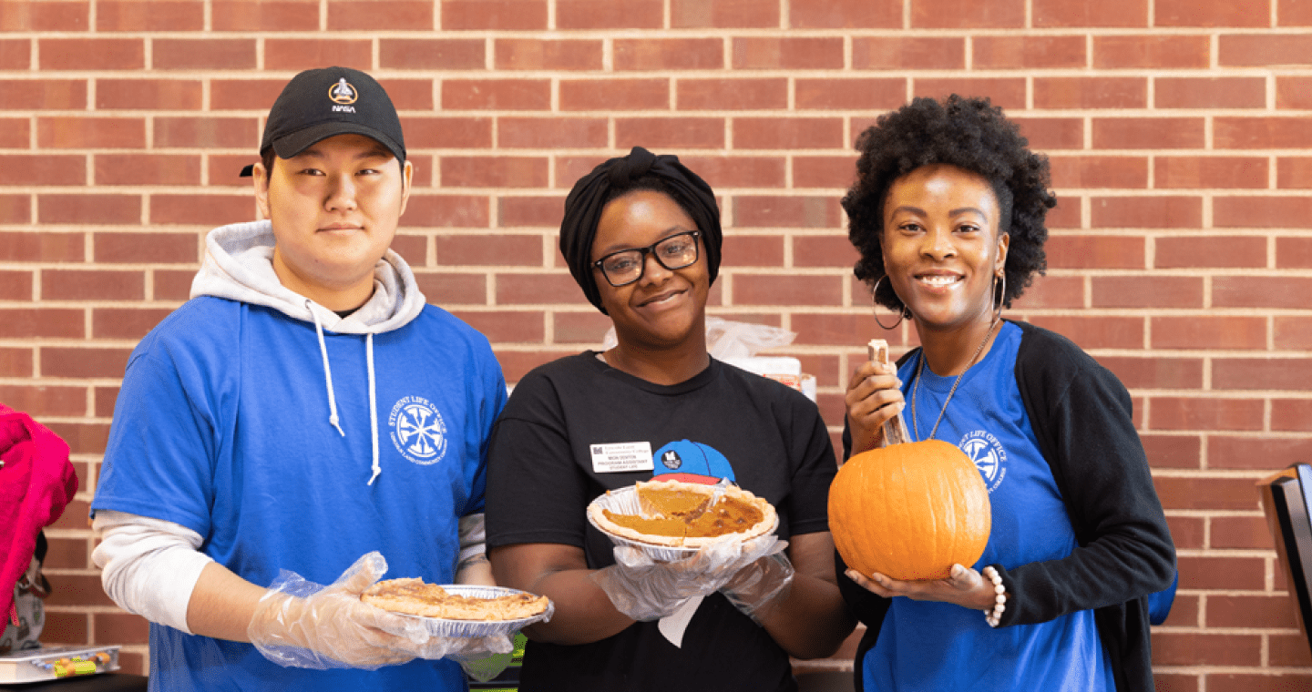 two students with LLCC employee holding pumpkin and pies smiling at the camera