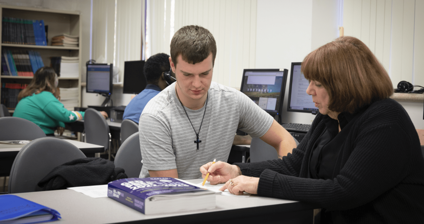 teacher instructing a student, with students working on computers in the background