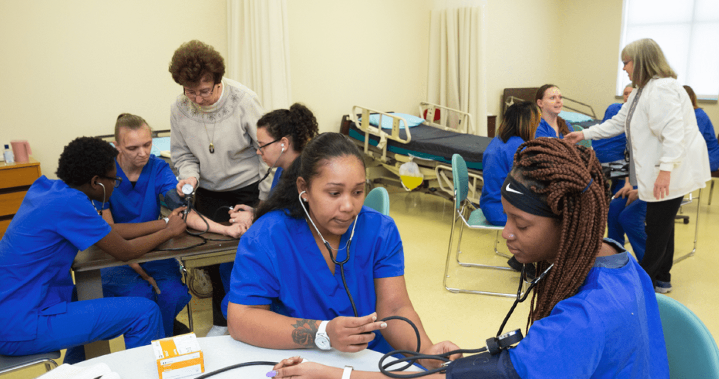 Nine students in scrubs learning from two instructors about how to take blood pressure readings