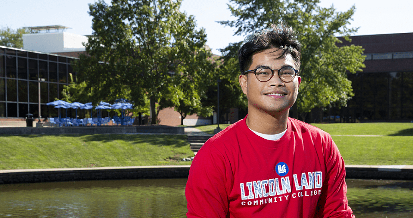 Student with LLCC shirt in foreground; lake and outdoor seating on campus in background