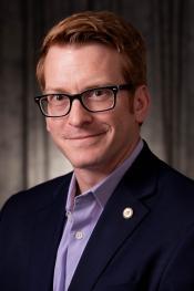 Headshot of Jacob K. Friefeld. He is a man wearing a navy suit with a purple button up shIrt and glasses.