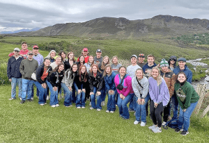 A group of college students poses for a group shot in a lush field in Ireland with a mountain in the background