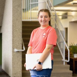 A young woman wearing a pink shirt and holding a laptop at her side.