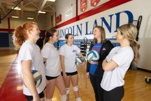 Volleyball players stand with their coach in a gym.