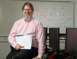 A man with gray-ish hair and glasses wearing a light-pink shirt sits for a photo in a classroom.