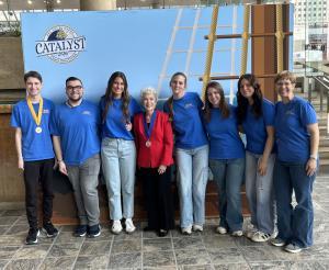 Nine people stand shoulder to shoulder in blue shirts; one wears a red blazer. Medals hang from necks. Backdrop reads “Catalyst 2026” with ship ropes.