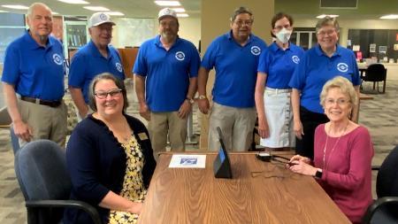 Members of the Springfield Sertoma Club wearing matching blue polo shirts stand in a row behind the dean of LLCC Library and LLCC's president, who are both seated at a table with a Portable InfoLoop device.