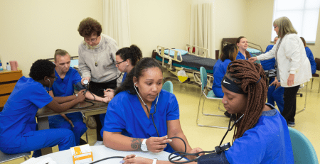 Nine students in scrubs learning from two instructors about how to take blood pressure readings