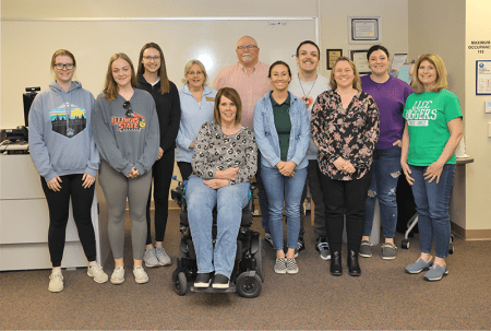 Gaye Catlin and her husband, Scott pose in a classroom with a group of Occupational Therapy Assistant students