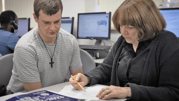 A student looks down at a piece of paper that a teacher is pointing to with a pencil.