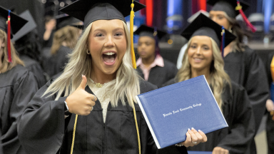 A female graduate smiles, giving a thumbs up with her right hand and holding her diploma cover in her left hand.