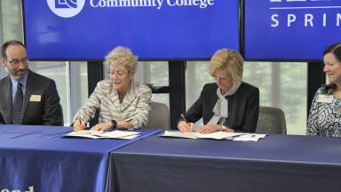 LLCC President Dr. Charlotte Warren and UIS Chancellor Dr. Janet Gooch sign agreement documents while seated at a table with other college and university officials.