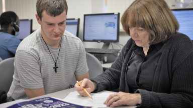 A student looks down at a piece of paper that a teacher is pointing to with a pencil.